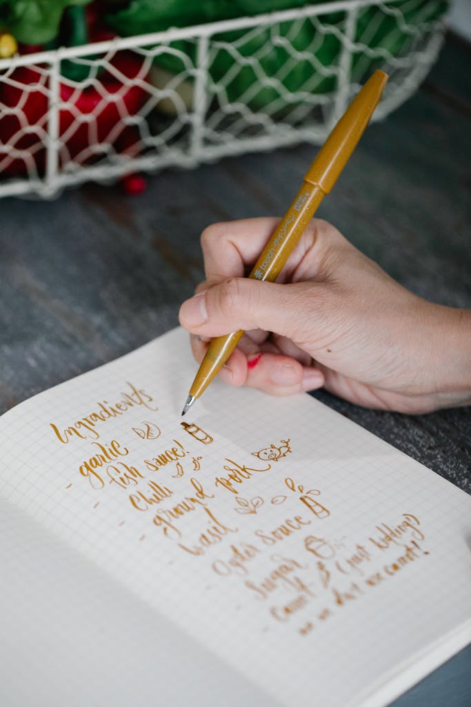 Photo by Katya Wolf Close-up of a hand writing ingredients list with a golden pen on a notepad.