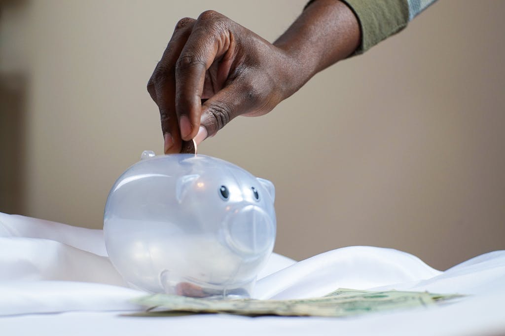 Photo by Joslyn Pickens Close-up of a person's hand placing coins into a transparent piggy bank to save money.